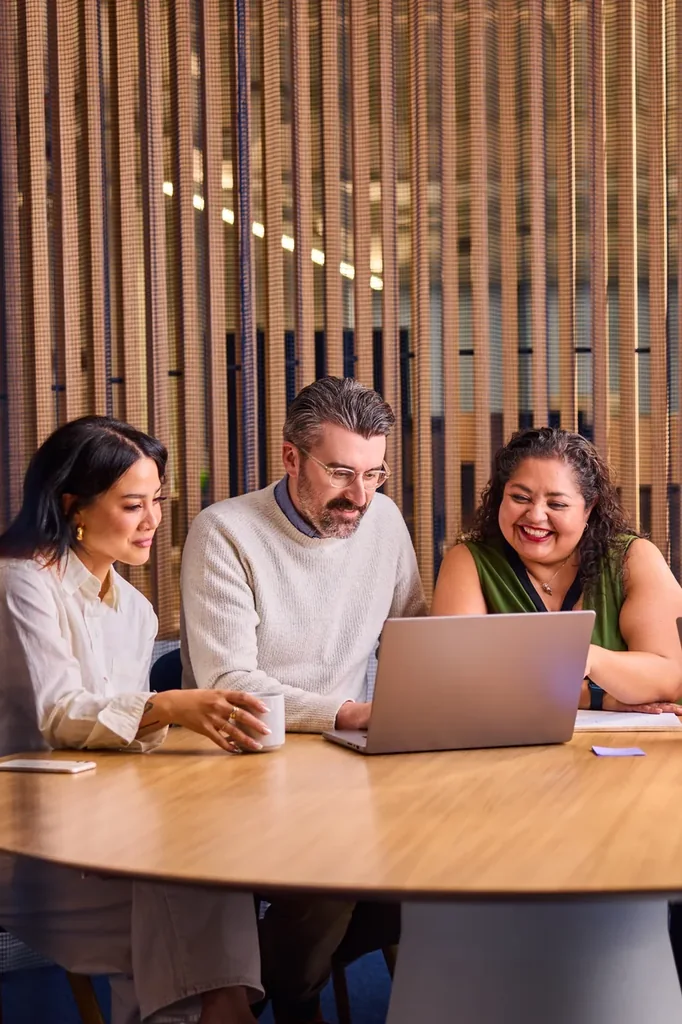 Tres profissionais colaborando em um notebook em mesa redonda de escritorio