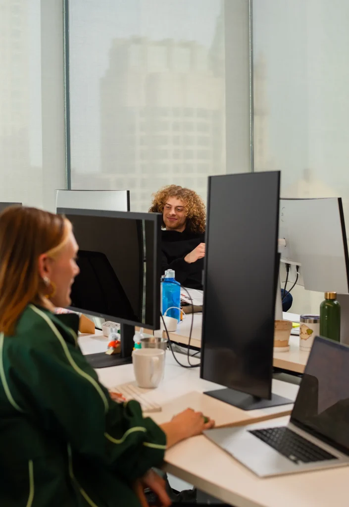 Desenvolvedor de cabelo cacheado sorrindo em conversa com colega de equipe, entre monitores de trabalho