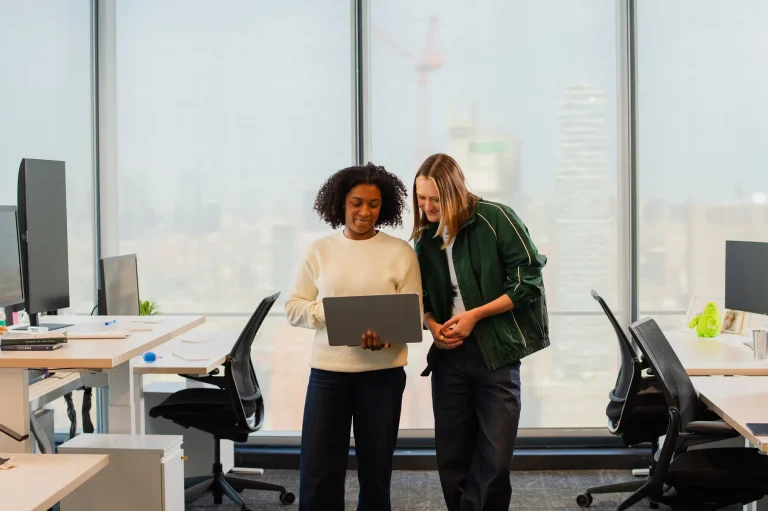 Duas profissionais revisando peça de campanha em laptop, com skyline ao fundo