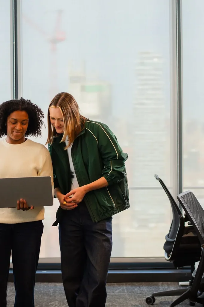 Duas profissionais conversando em pe com notebook em escritorio com vista da cidade